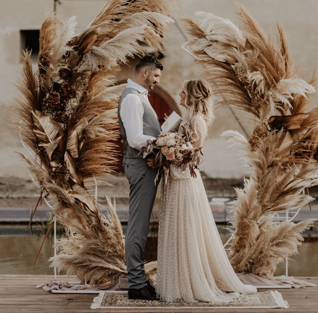 Ein glückliches Brautpaar hält sich, vor einem Trau bogen im Boho-Stil fest in den Armen. Durch die runde Komposition des Pampasgrases wirkt der Bogen wie ein Ring der Ewigkeit.